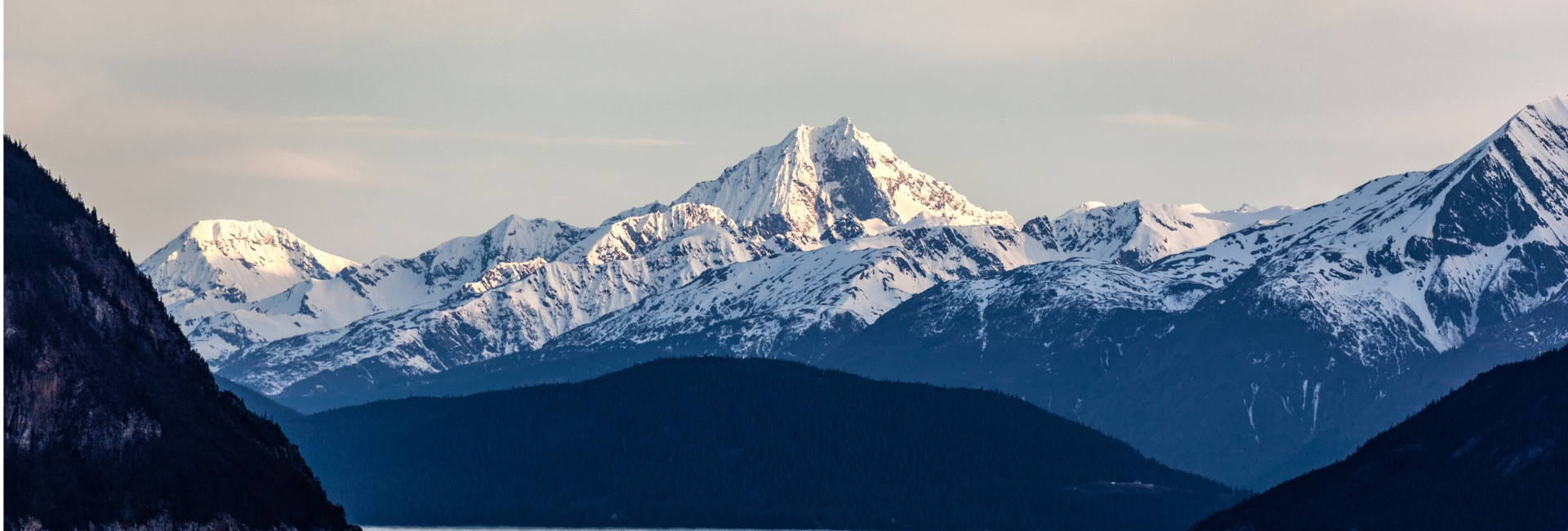 30+ Years of Adventure 11 - Skagway Excursions - Skagway Excursions Snow-covered mountain range with sunlit peak, dark forested foothills, and a calm lake in the foreground.